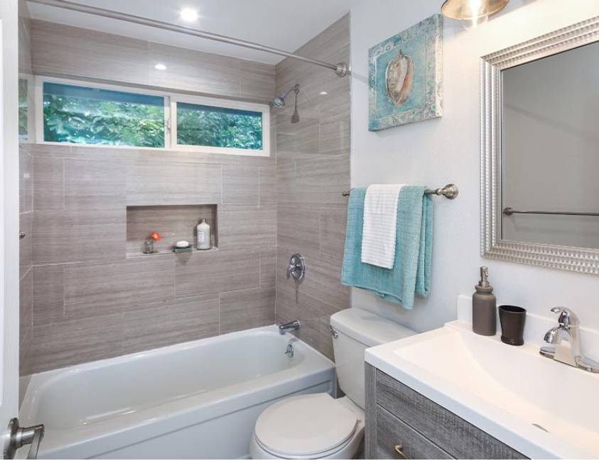 Modern bathroom with beige tiles, bathtub, and window with greenery view